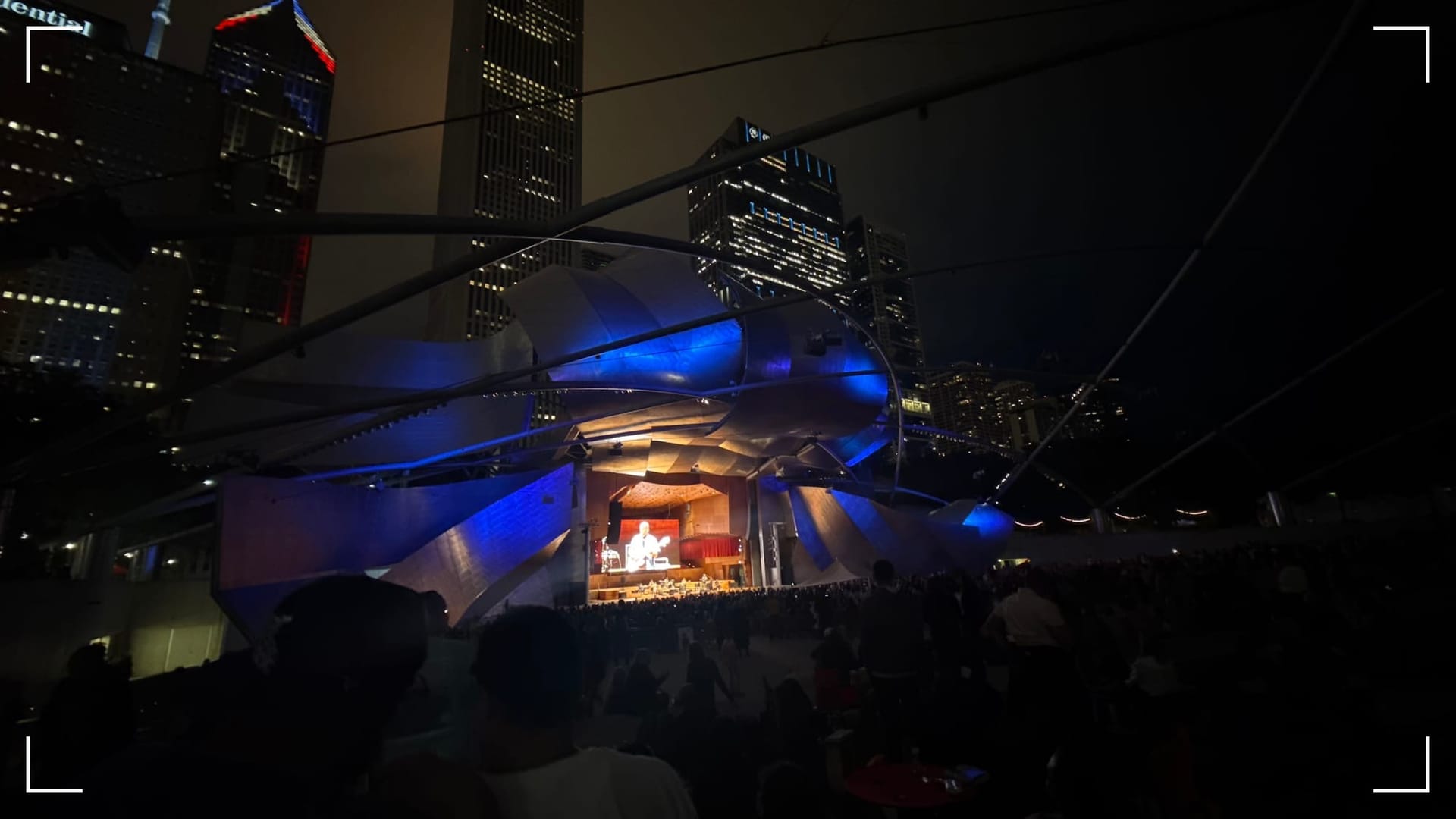 The Pritzker Bandshell/Pavilion in Chicago's Millennium Park, lit in blue at night against an overcast, hazy Chicago backdrop at night.